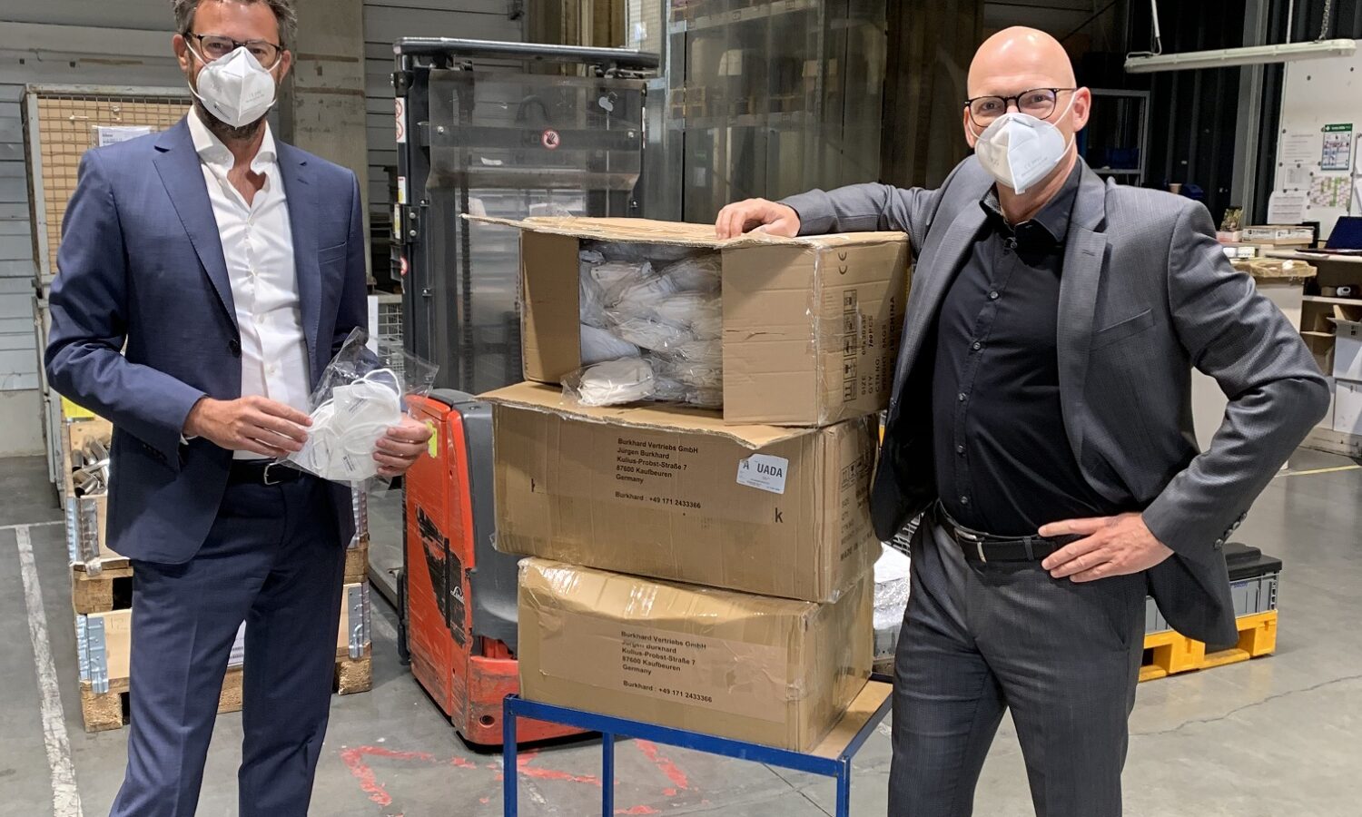 Zwei Männer mit Schutzmasken stehen neben Kartons mit Masken in Produktionshalle