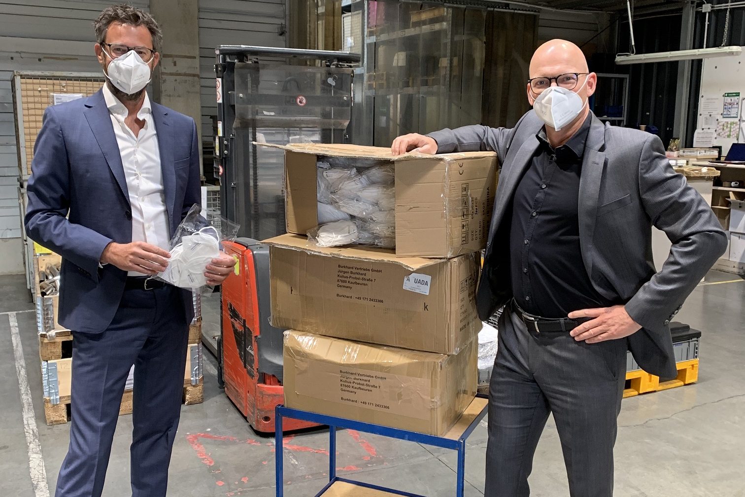 Masken_Erste_Lieferung_klein Zwei Männer mit Schutzmasken stehen neben Kartons mit Masken in Produktionshalle