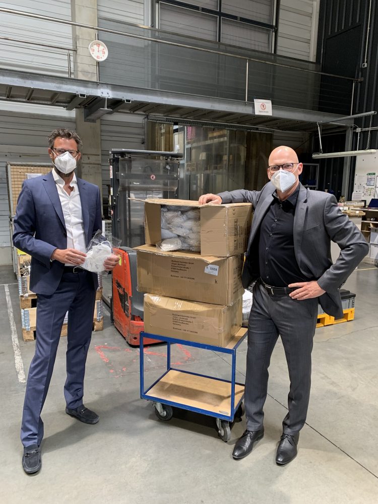 Zwei Männer mit Schutzmasken stehen neben Kartons mit Masken in Produktionshalle