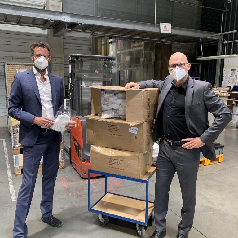 Zwei Männer mit Schutzmasken stehen neben Kartons mit Masken in Produktionshalle