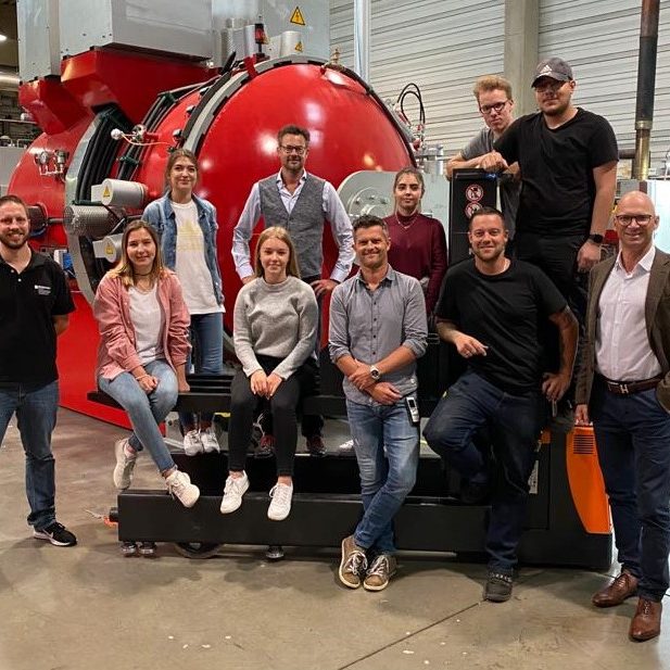 Teamgruppe steht vor großer roter Industrieanlage in Produktionshalle
