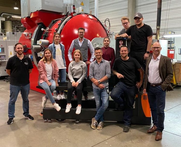 Teamgruppe steht vor großer roter Industrieanlage in Produktionshalle