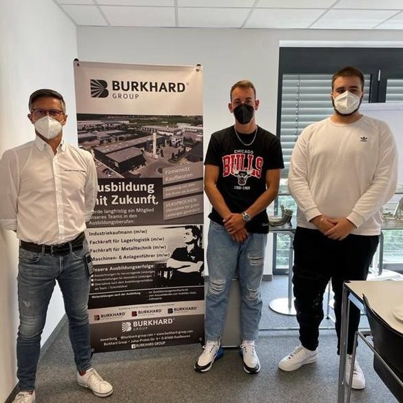 Drei Männer mit Mundschutz stehen neben BURKHARD Ausbildungs-Rollup im Büro