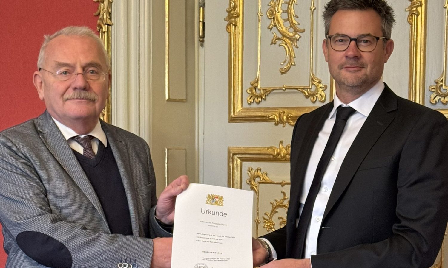 Zwei Männer überreichen eine Urkunde vor einer verzierten Tür im Landgericht